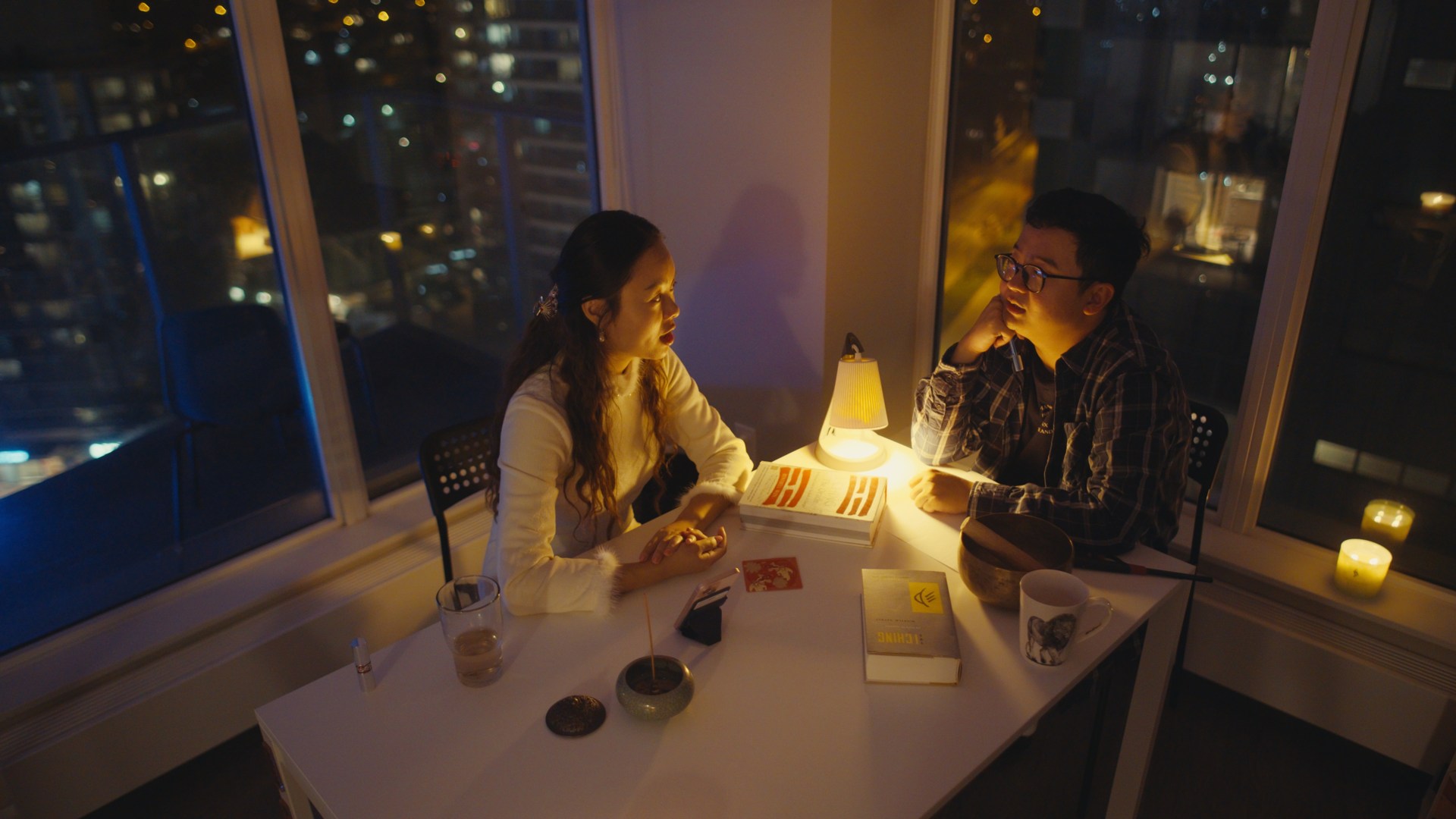 Two people talking at a lamp-lit table overlooking the city at night
